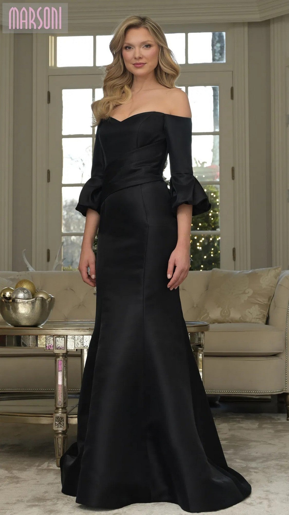 A woman poses indoors before a sofa and coffee table, wearing the black Marsoni MV1355 long formal dress with off-the-shoulder flared sleeves and a mermaid silhouette. Large windows and a holiday tree are in the background.